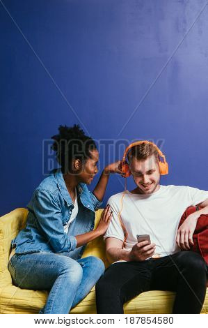 Young interracial couple in quarrel at home, white man ignore black woman. Wear headphones and look at phone, do not listen, relationship problem, inattentive concept.