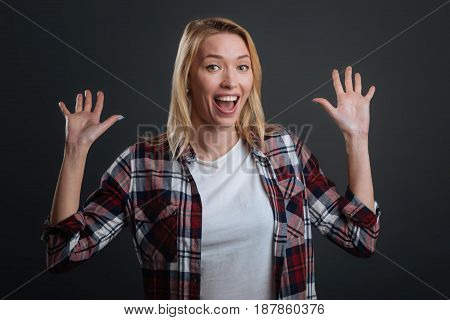 What a surprise. Expressive gorgeous young lady throwing her hands up in excitement while being very emotional and standing isolated on grey background