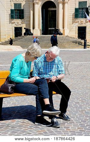 VALLETTA, MALTA - MARCH 30, 2017 - Couple sitting on a bench reading a guide book with the Auberge de Castille to the rear in Castille Square Valletta Malta Europe, March 30, 2017.