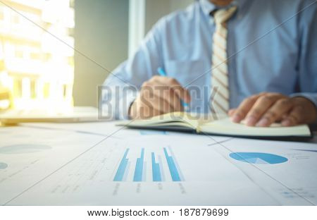 Young man working and writing in Book on his desk with financial grap document business person at workplace.