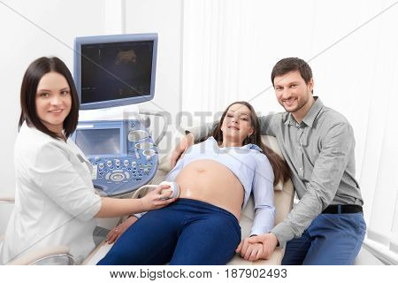 Female doctor screening stomach of pregnant woman using special tools. Lovely husband carrying about wife on procedure of ultrasound in hospital and embracing woman. Nurse smiling at camera.