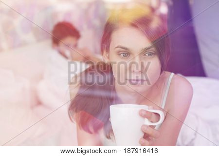 Thougtful woman having coffee at home with man lying in background