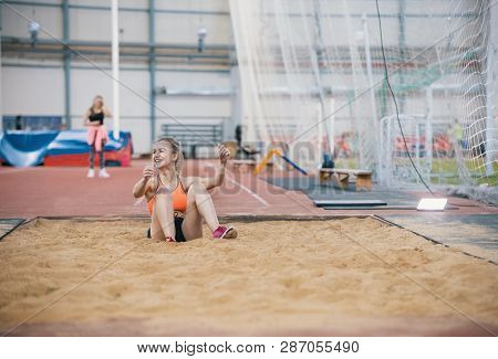 Young Blonde Smiling Women Performing A Long Jump In The Sports Arena. Sitting In The Sand