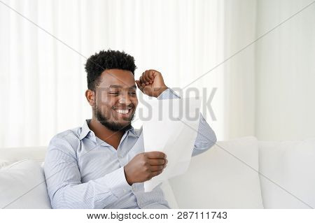 The African Male Reads A Letter With Good News Sitting On The Couch At Home. An Euphoric Male Is Hap
