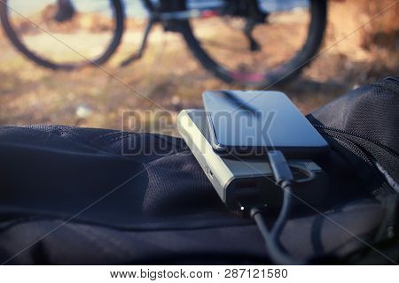Portable Charger Charges The Smartphone. Power Bank With Cable Against The Background Of Bicycle.