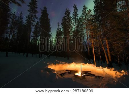 A Fireplace In Winter Forest With Aurora Borealis (northern Light) Shining On Sky Behind Trees In No