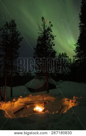Fireplace With A Wooden Cottage And Trees Covered In Snow Behind And Aurora Borealis (northern Light