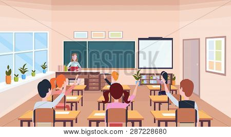 Pupils Raising Hands To Answer Question Teacher Sitting Desk Education Concept Modern School Classro