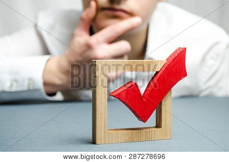 A Man Holds His Hand At The Chin And Assesses The Red Tick For A Vote. Assessing The Transparency Of