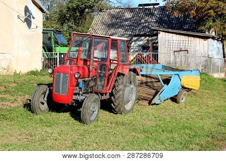 Red Tractor Parked Next To Family House With Old Agricultural Equipment In Background Surrounded Wit