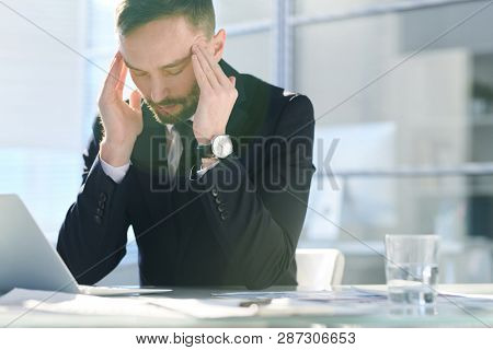 Tired man in formalwear touching his temples while sitting by desk in front of laptop while trying to concentrate