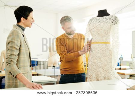 Contemporary mature tailor or fashion designer measuring waist girth of white lace dress on dummy with his client near by