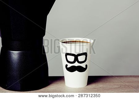 closeup of coffeemaker and a disposable cup with coffee, with a pair of glasses and a mustache drawn in it, depicting a man face, on a wooden table