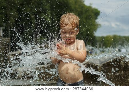 Funny Baby Boy Trying To Cauch Water Stream In Fountain. Cute Toddler Playing In The City Fountain