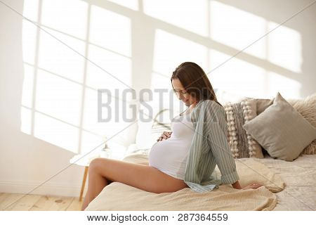 Sideways Shot Of Attractive Young Brunette Female In Last Month Of Pregnancy Sitting On Bed In Brigh