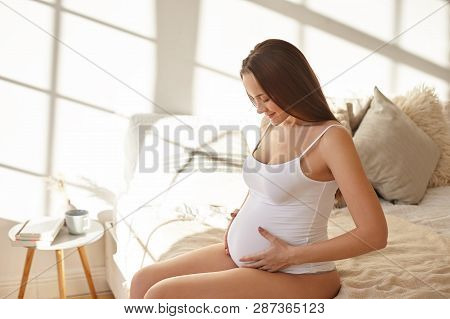 Amazing Young Pregnant Female Wearing Only White Tank Top Sitting In Bedroom After Awakening, Feelin