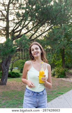 Cute Girl Is Having A Walk In The Park Holding A Bottle Of Orange Juice In A Sunny Wam Day