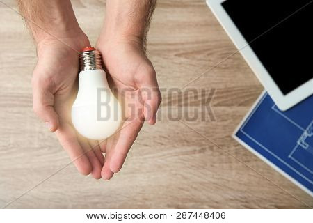 Man Holding Lamp Bulb Over Table With Tablet And Blueprint, Top View. Space For Text