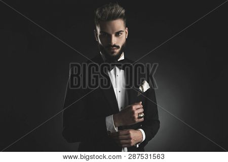 man in tuxedo with bowtie and white hankerchief fixing his sleeve on black background