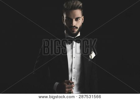 man in tuxedo with bowtie and white hankerchief adjusting his lounge jacket on black background