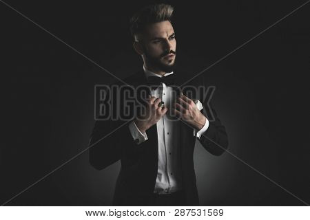 man in tuxedo with bowtie and white hankerchief adjusting his collar while looking away on black background