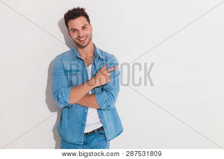 Attractive man smiling and holding his hands crossed and pointing fingr to side  while looking at the camera and standing against a white wall