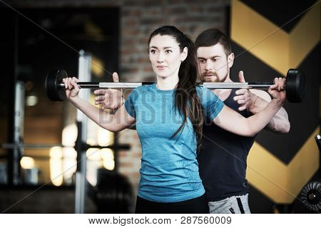 Personal trainer man helps woman work with barbell at squat exercises in gym