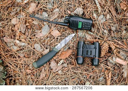 Binoculars, Radio Set And Hunting Knife On The Ground Covered With Pine Needles