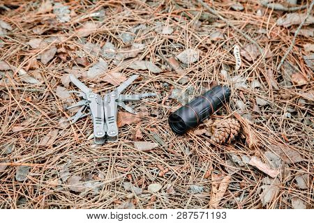 Monocle And Multitool Knife On The Ground Covered With Pine Needles