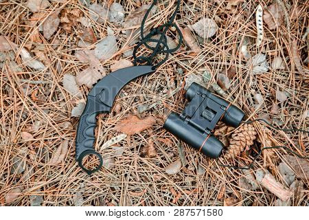 Binoculars And Karambit Knife On The Ground Covered With Pine Needles