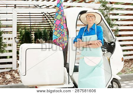 Waist Up Portrait Of Senior Woman Posing In Garden Standing Next To Moving Cart With Arms Crossed, C