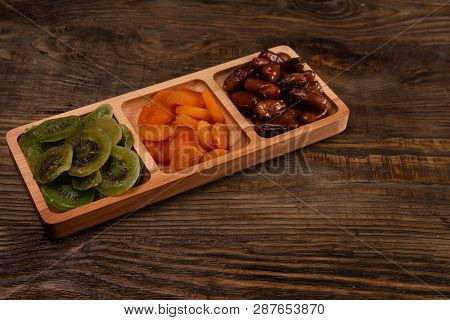 Dates, Dried Apricots And Kiwis In A Compartmental Dish On A Dark Wooden Table.
