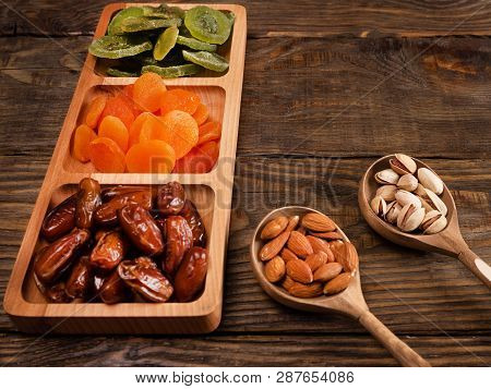 Dates, Dried Apricots And Kiwis In A Compartmental Dish And Nuts In A Wooden Spoon On A Dark Wooden 