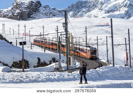 Zermatt, Switzerland - April 12, 2017: The Red Gornergrat Train And A Man Skiing On A Piste Next To 