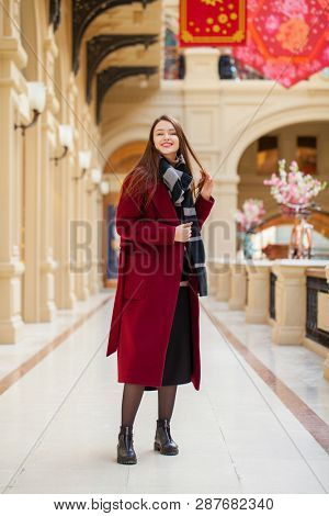 Full body portrait. Young beautiful woman in stylish in a long burgundy coat, indoor shop