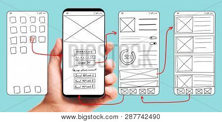 UI development. Male hand holding smartphone with wireframed user interface screen prototypes of a mobile application on blue background.
