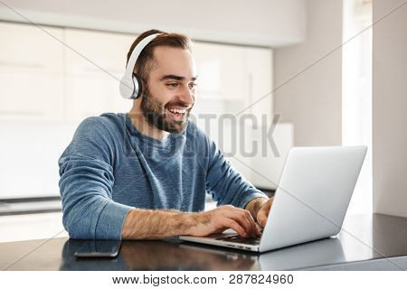Happy handsome man freelancer sitting at the kitchen table, working on laptop computer, wearing headhones