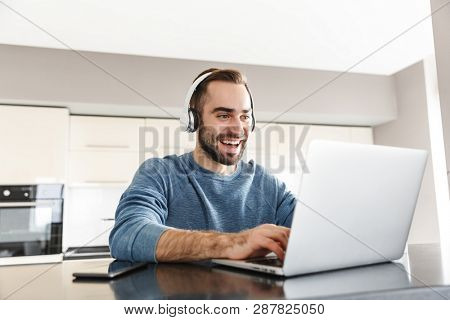 Happy handsome man freelancer sitting at the kitchen table, working on laptop computer, wearing headhones