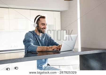 Happy handsome man freelancer sitting at the kitchen table, working on laptop computer, wearing headhones