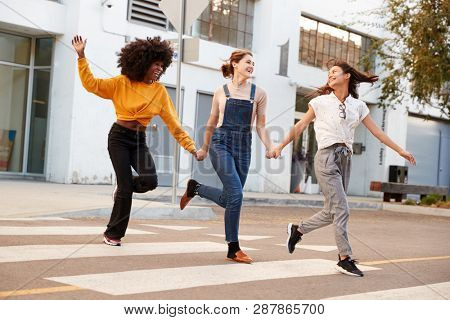 Three millennial hip girlfriends holding hands and laughing as they run across a pedestrian crossing
