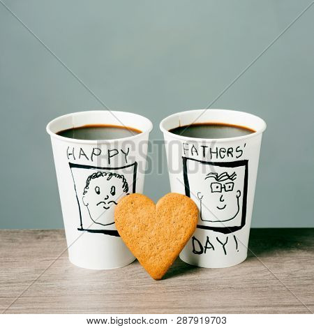 closeup of two disposable cups with coffee, decorated with the drawing of two men and the text happy fathers day handwritten in it, and a heart-shaped cookie on a wooden table