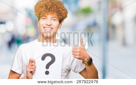 Young handsome man holding paper with question mark happy with big smile doing ok sign, thumb up with fingers, excellent sign