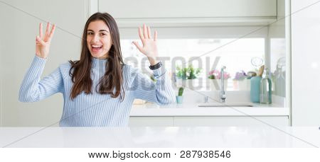 Wide angle picture of beautiful young woman sitting on white table at home showing and pointing up with fingers number eight while smiling confident and happy.