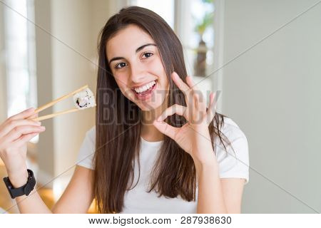 Beautiful young woman eating asian sushi using chopsticks doing ok sign with fingers, excellent symbol