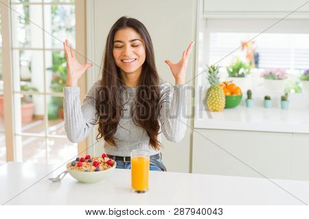 Young woman eating healthy breakfast in the morning celebrating mad and crazy for success with arms raised and closed eyes screaming excited. Winner concept