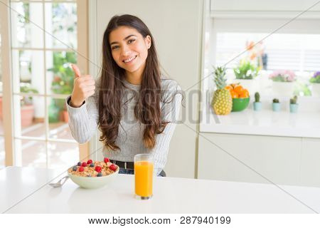Young woman eating healthy breakfast in the morning doing happy thumbs up gesture with hand. Approving expression looking at the camera with showing success.