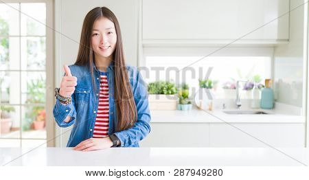Beautiful Asian woman wearing denim jacket on white table doing happy thumbs up gesture with hand. Approving expression looking at the camera showing success.
