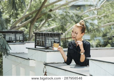 Pensive Business Lady Biting Side Piece Of Her Glasses When Drinking Coffee In Outdoor Cafe