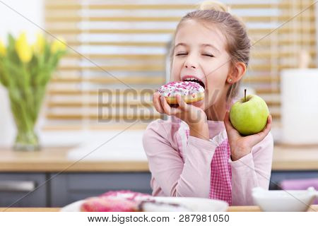 Cute little girl choosing between apple and donut in the kitchen