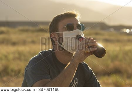 A Young Man, With Glasses, Tries To Drink A Beer Wearing A Protective Face Mask.